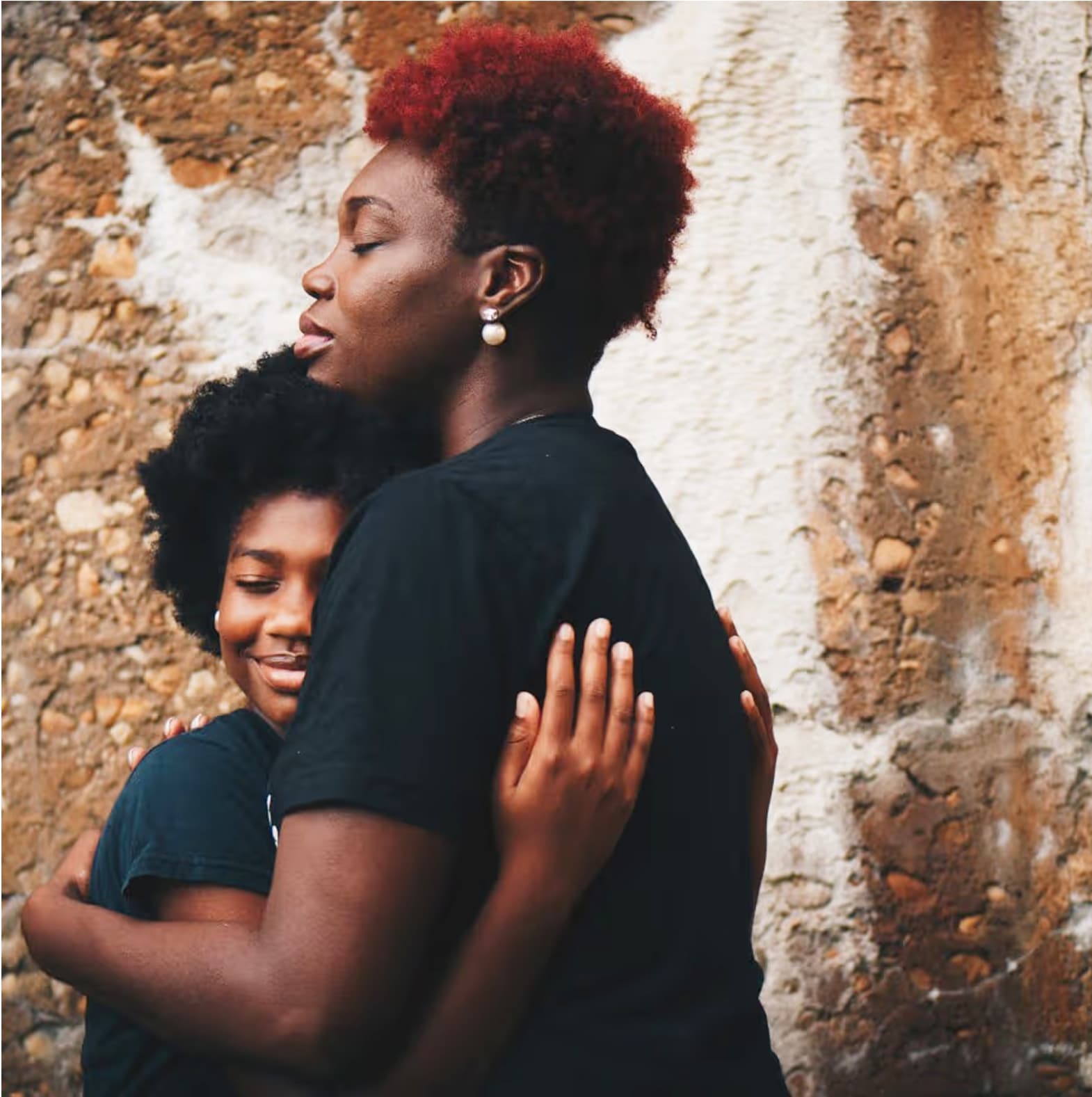 A tender moment captured in profile showing two people embracing, wearing black t-shirts against a textured stone wall background. One has vibrant red-tinted natural hair styled in a short cut, while the other has dark natural curls. Their peaceful expressions convey warmth and connection.