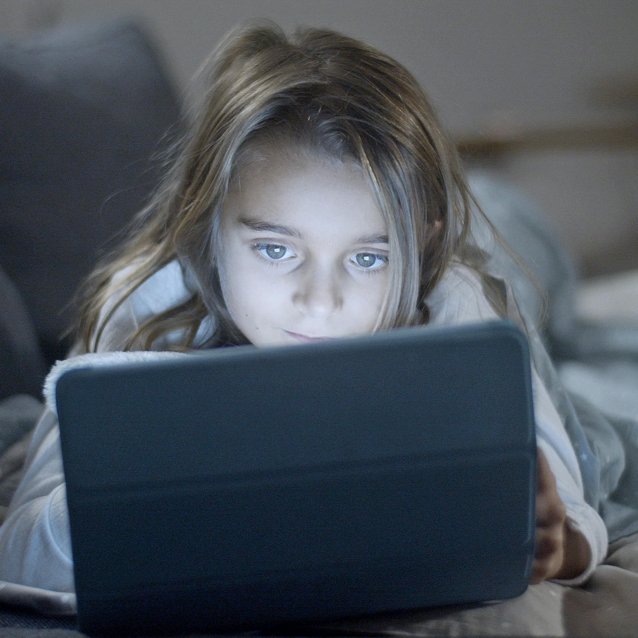 Young girl laying on her stomach in a dimly-lit room looking at an iPad screen