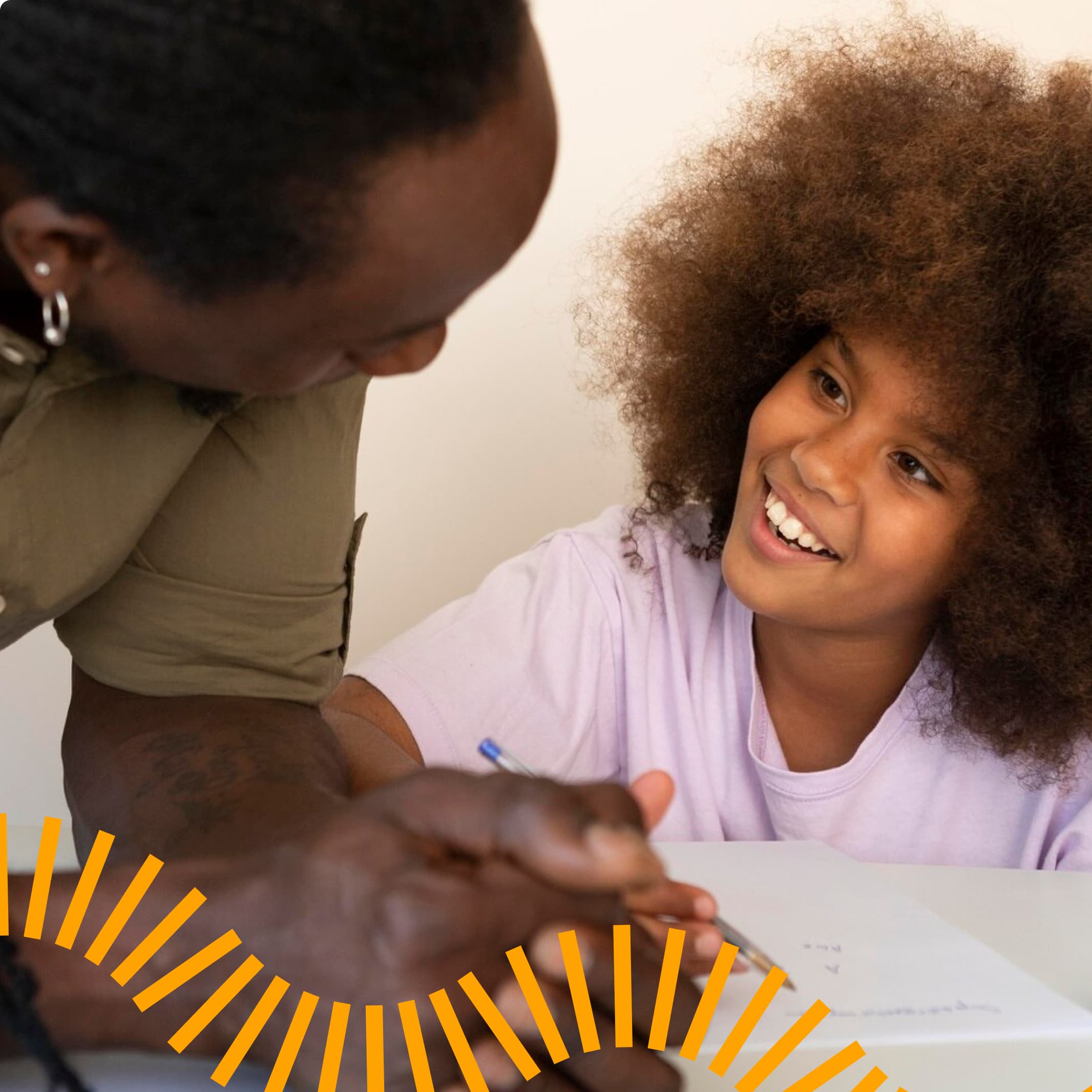 An engaging tutoring moment captured as a student with natural curly hair in a light purple t-shirt smiles brightly while receiving academic guidance. The tutor, wearing an olive-colored shirt, leans in to assist with written work on the desk.