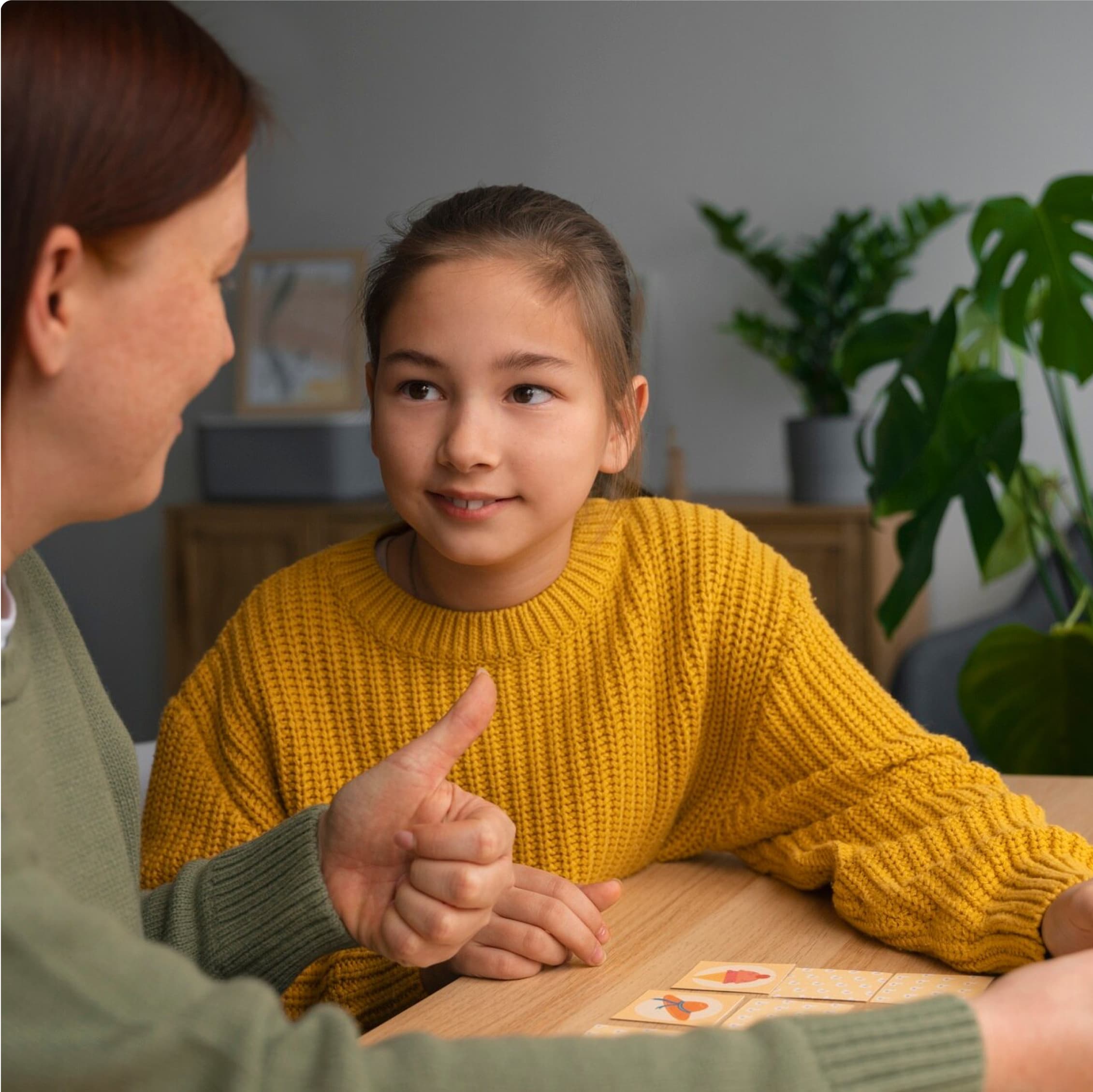 A heartwarming scene of interaction between a parent and child at a wooden table, with the young person wearing a bright yellow knit sweater engaging in what appears to be an educational card game. Indoor setting with houseplants in the background.