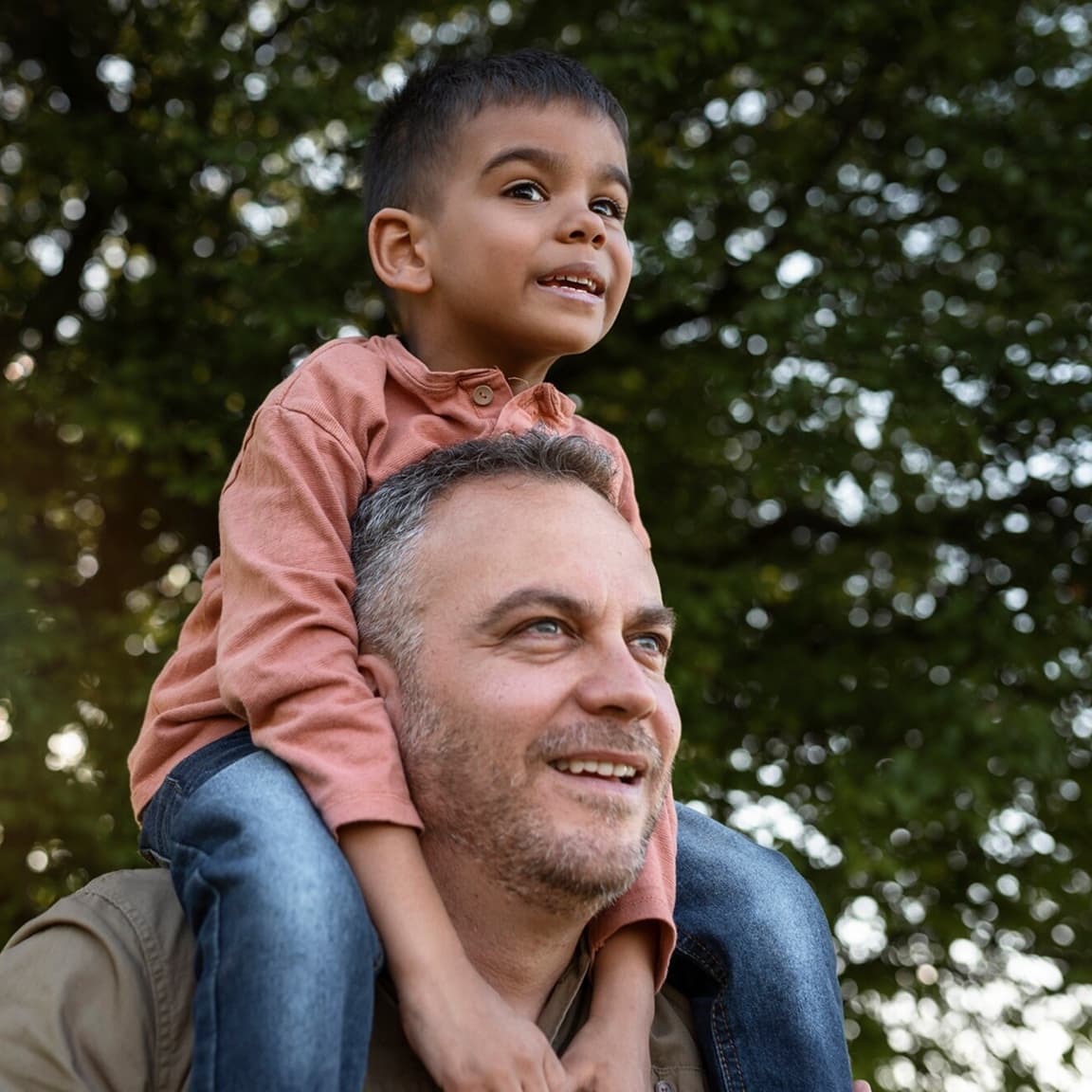 A heartwarming outdoor scene showing a young child in a coral-colored shirt sitting on the shoulders of an adult with salt-and-pepper hair, both smiling brightly against a blurred green foliage background