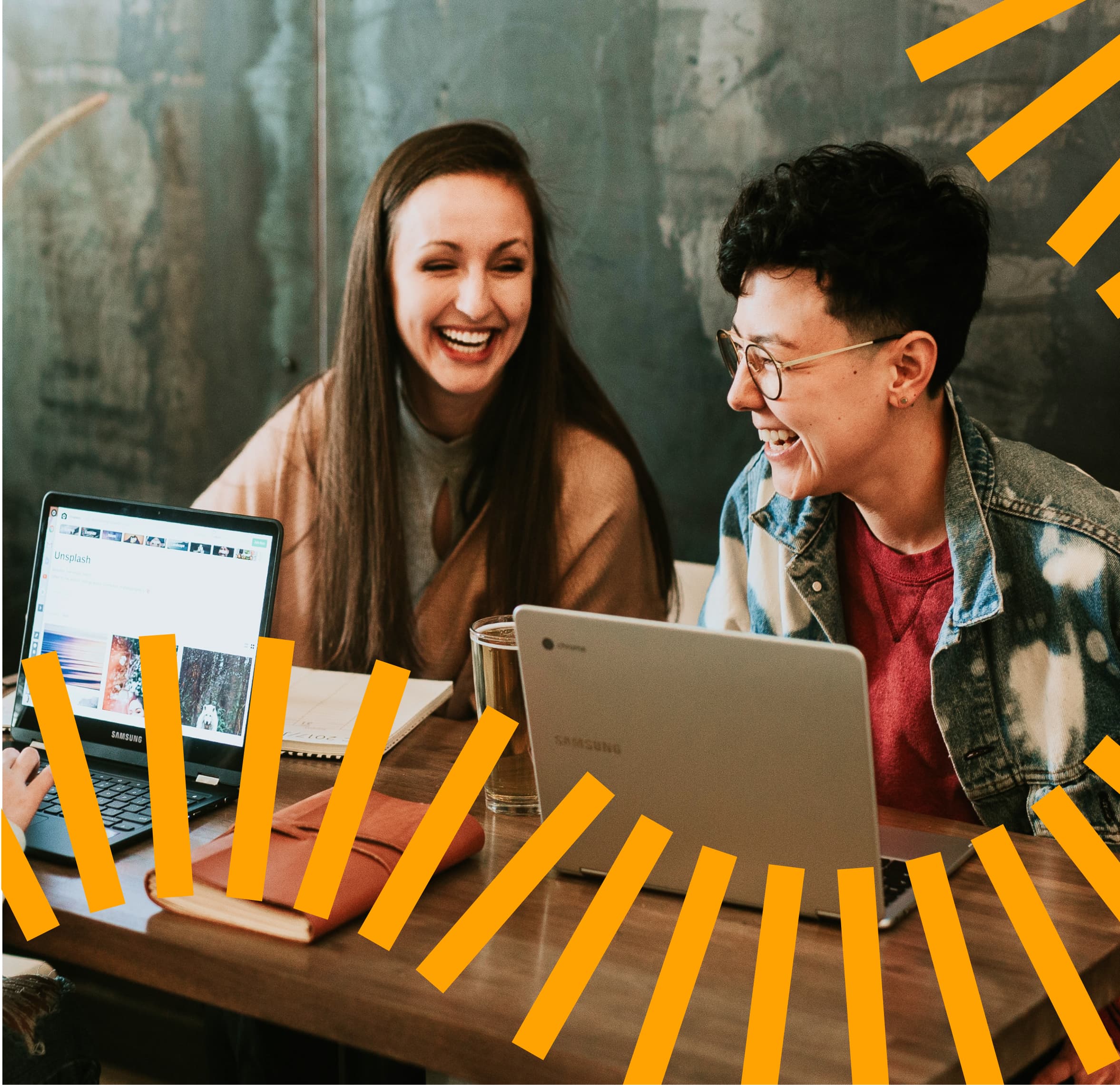Two people sharing a lighthearted moment while working together at a wooden table with laptops. One person wears a beige sweater while the other sports a denim jacket and glasses. The scene is decorated with stylized orange geometric lines and set against an industrial-style wall background.