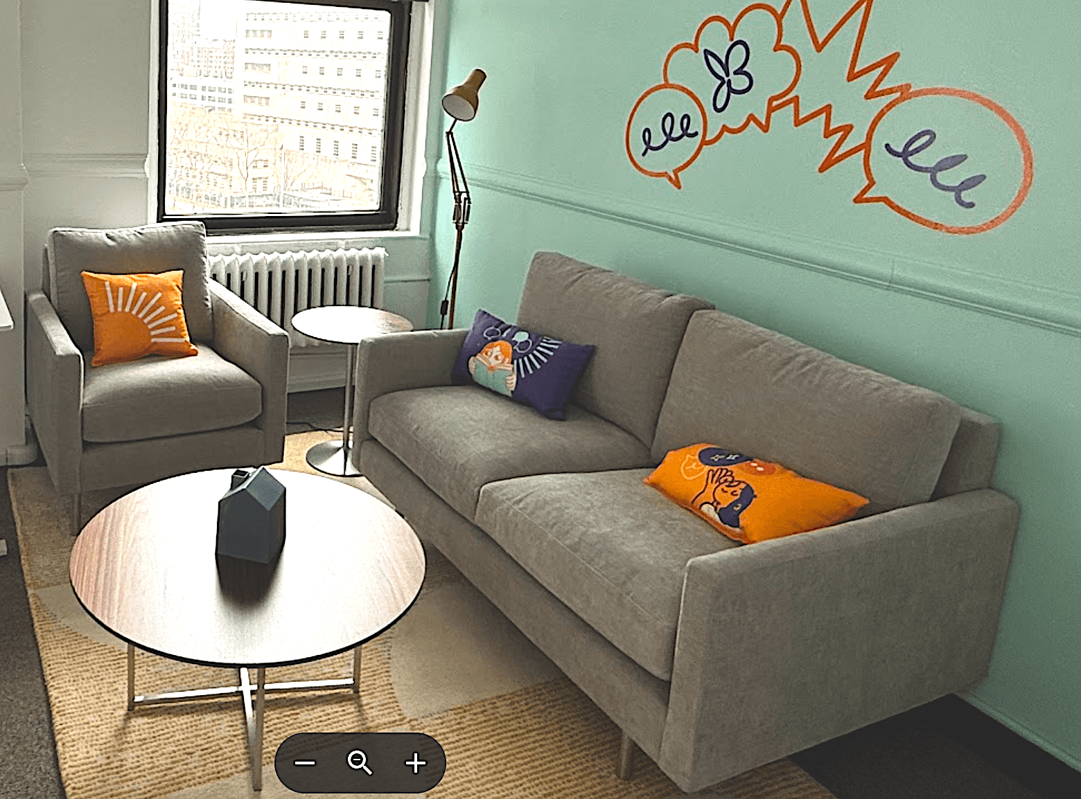 A small living space with a sofa with colorful pillows, chair with an orange pillow, and coffee table, green wall with illustrations on it