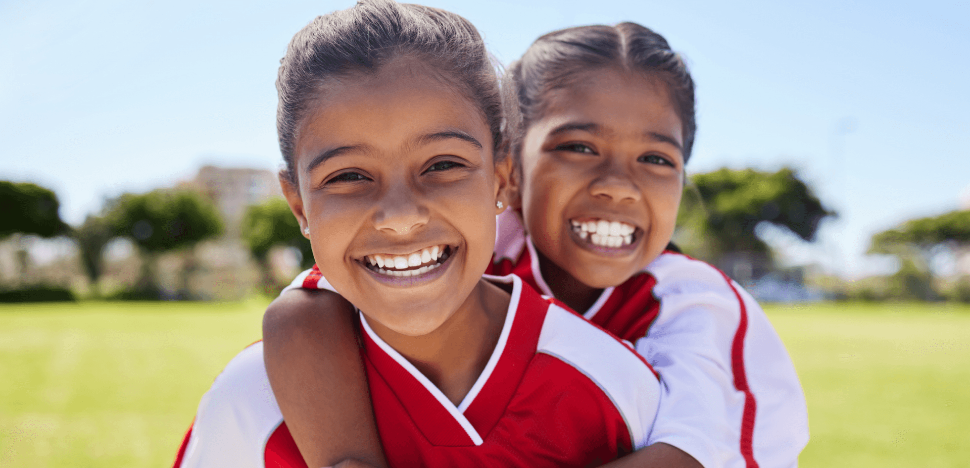 Two smiling girls in soccer uniforms on a grassy field, one hanging over the shoulders of the other