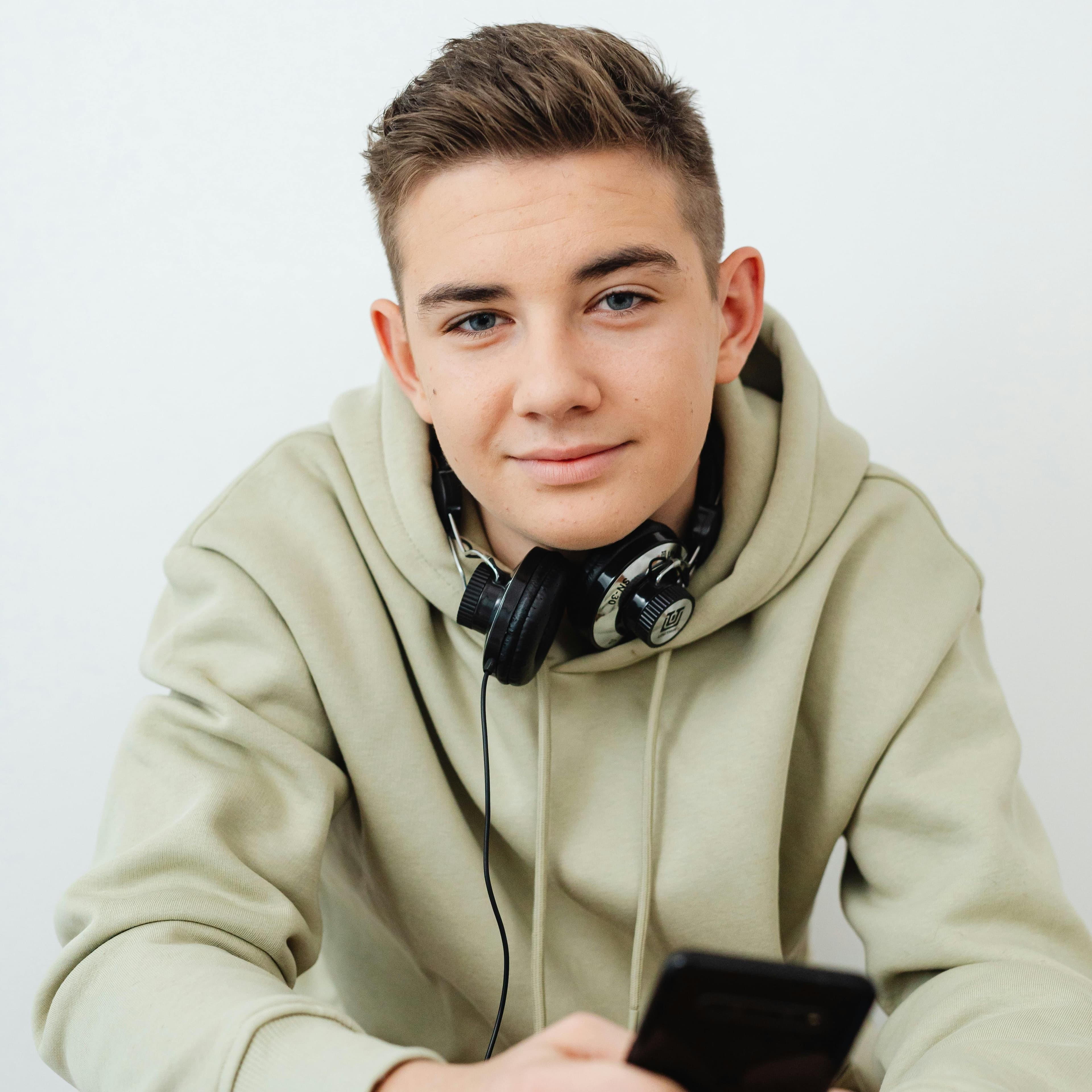 Young man in hoodie with headphones around his neck holding a phone and looking at camera