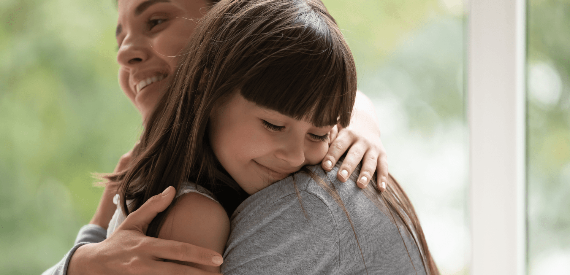 Adult hugging and comforting a young girl both standing in front of a big window