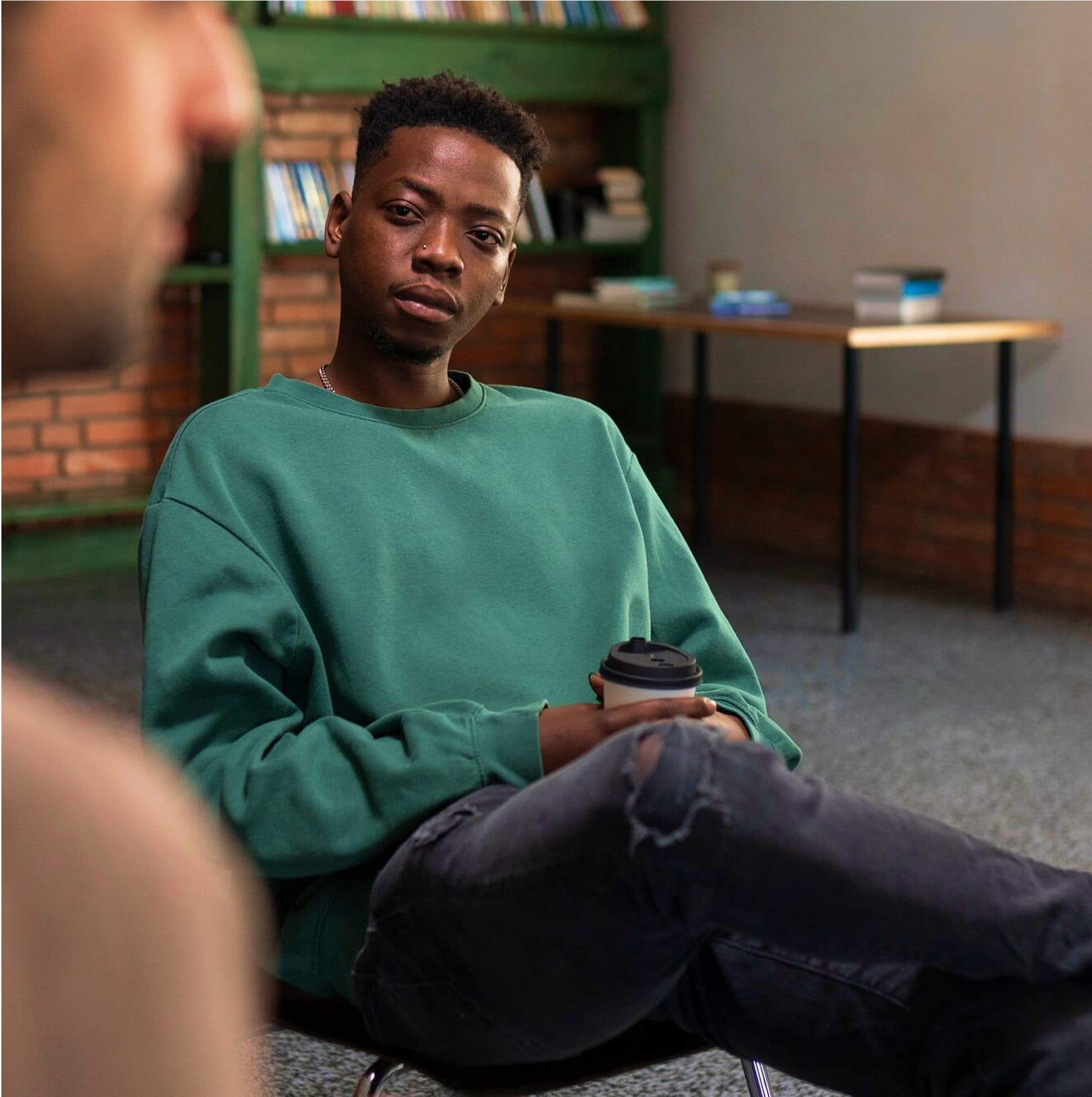 A person wearing a green sweatshirt sits casually in what appears to be an office or study space, holding a takeout coffee cup. The background shows a green bookshelf and brick wall, creating a modern yet cozy atmosphere