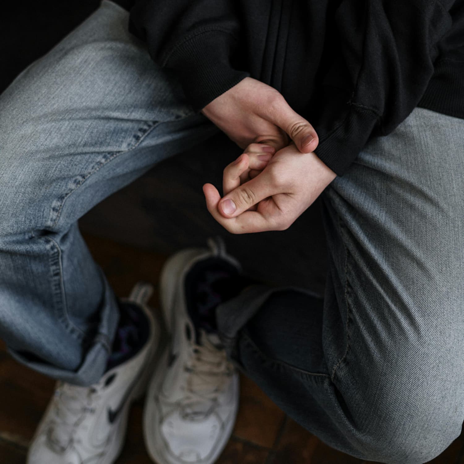 Teen sitting with view of their lap from above, they are fidgeting with their sweater sleeve, wearing jeans, and tennis shoes