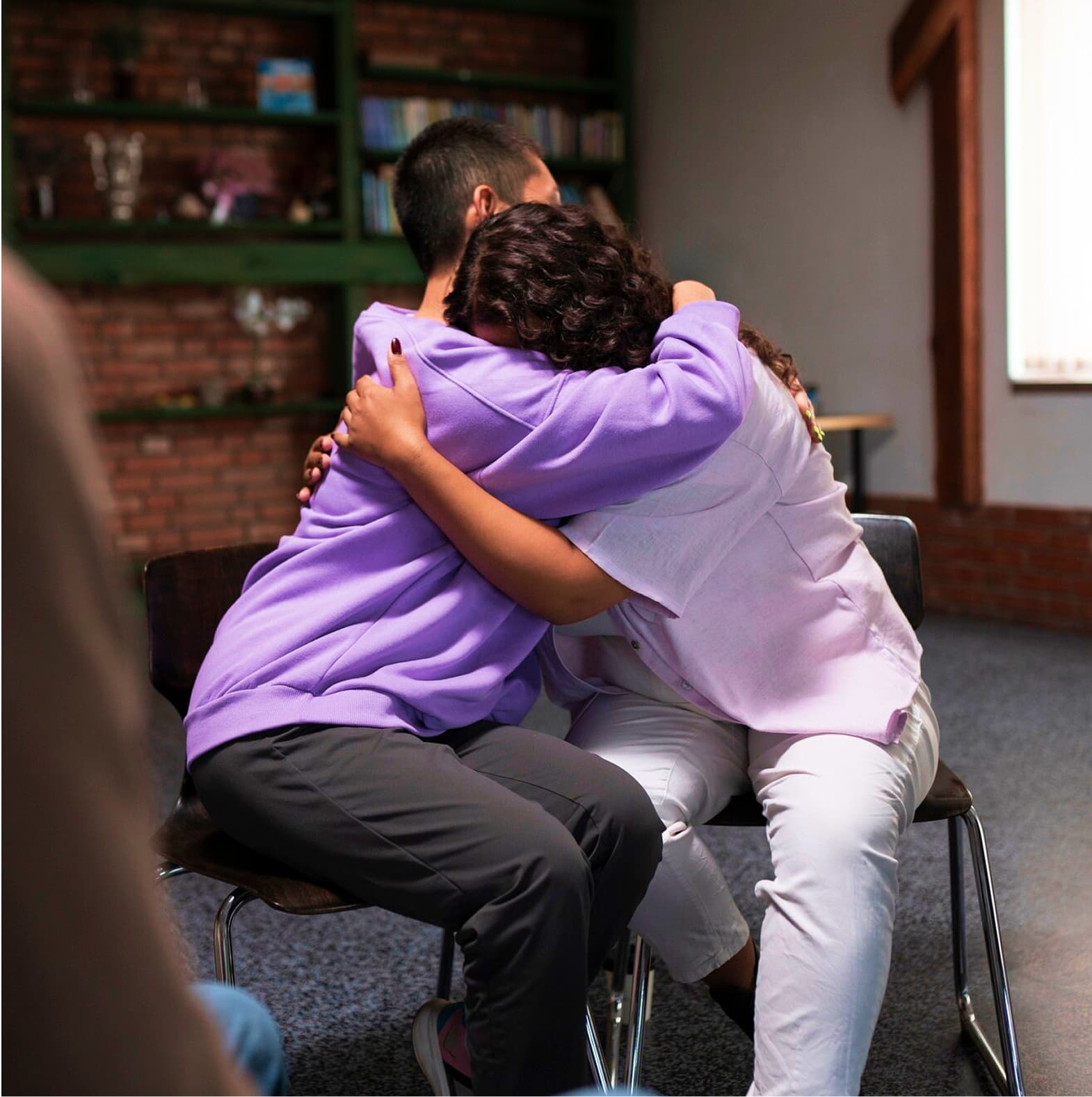 Two people sharing an emotional side-hug while seated on chairs in what appears to be a therapy or support group setting, with one person wearing a purple sweatshirt and the other in white, against a brick wall background with bookshelves
