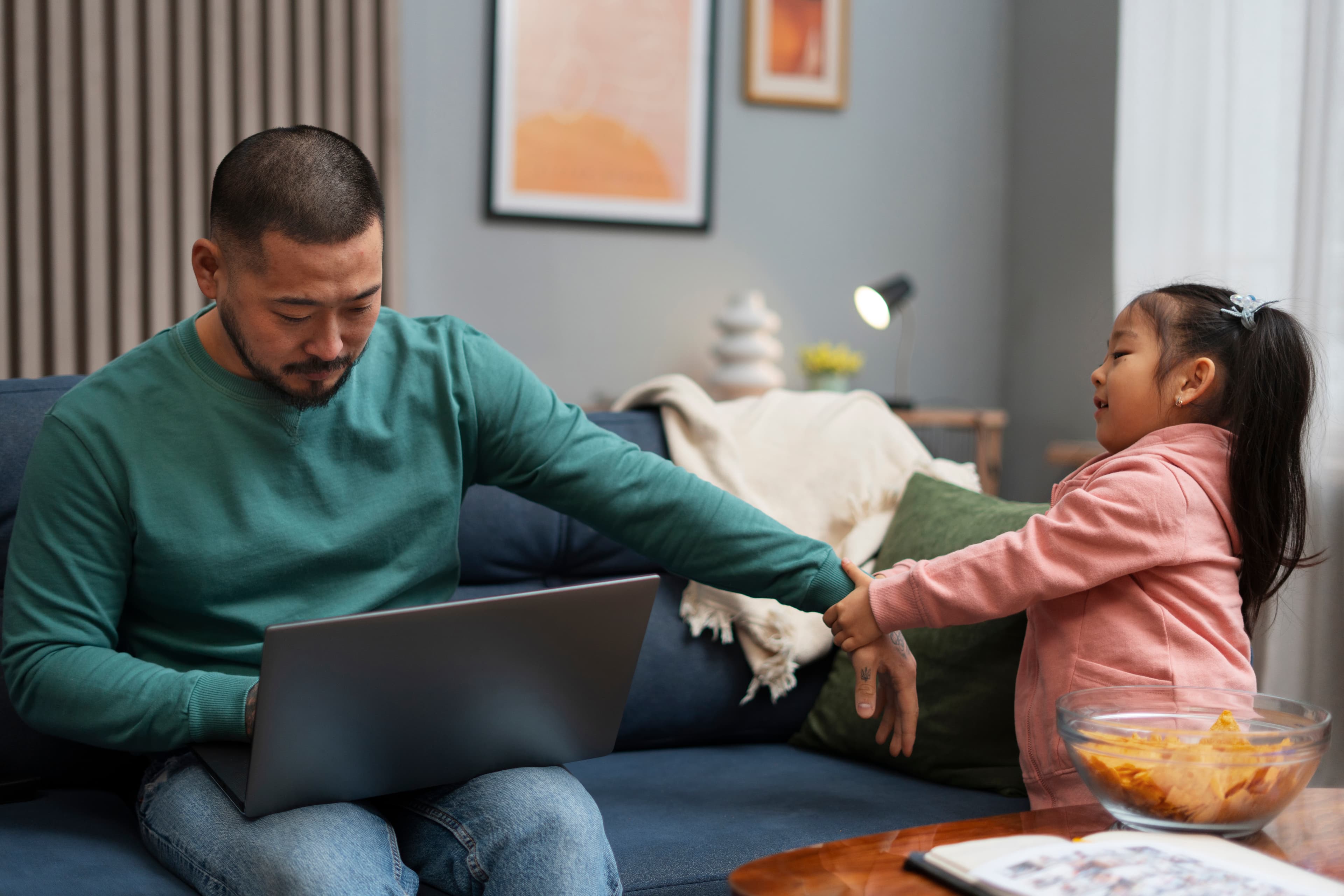 Man sitting on couch working on computer while little girl pulls on his arm to get his attention