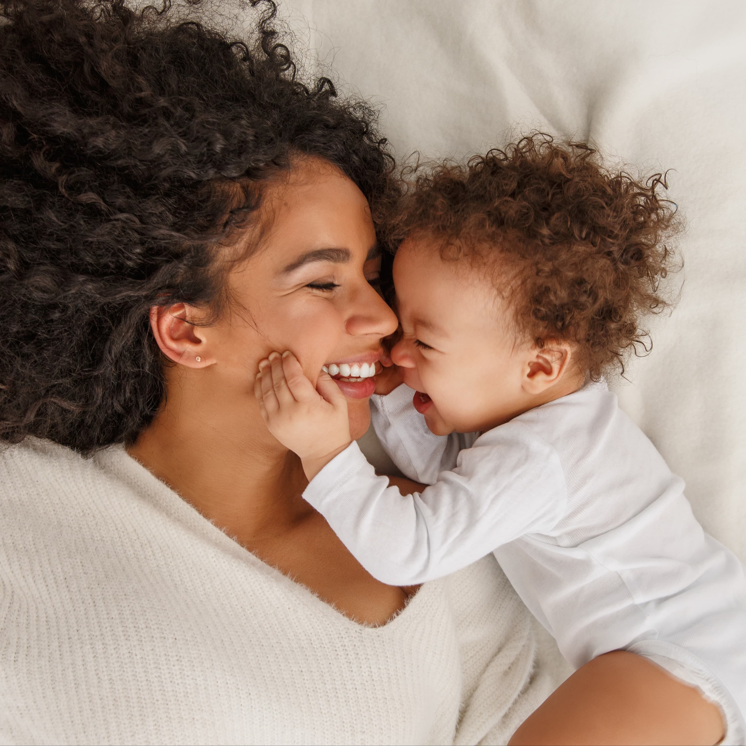 Mom laying on her back looking at her baby and smiling, baby is on their side smiling and reaching to touch mom's face